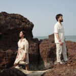 A romantic couple in a pre-wedding photoshoot on a South Goa beach during golden hour. They are dressed in flowing boho-chic outfits, with the woman in a pastel floral dress and the man in light casual wear. The vibrant scene features soft waves, swaying palm trees, and a sunset casting golden, pink, and purple hues. Subtle hints of South Goa landmarks, like Cabo de Rama Fort, appear in the background, creating a dreamy and serene atmosphere.