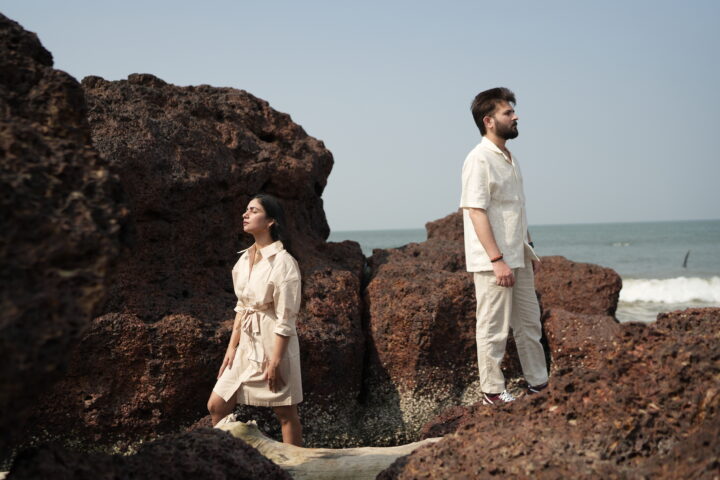 A romantic couple in a pre-wedding photoshoot on a South Goa beach during golden hour. They are dressed in flowing boho-chic outfits, with the woman in a pastel floral dress and the man in light casual wear. The vibrant scene features soft waves, swaying palm trees, and a sunset casting golden, pink, and purple hues. Subtle hints of South Goa landmarks, like Cabo de Rama Fort, appear in the background, creating a dreamy and serene atmosphere.